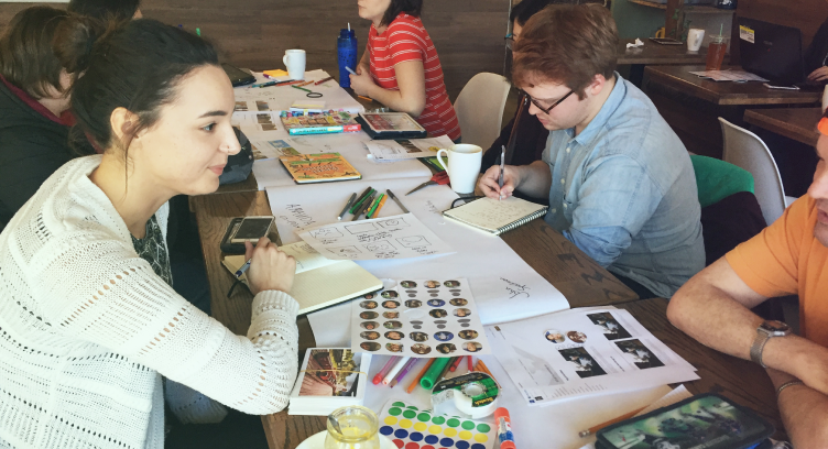 A group of people sit on either side of a long cafe table covered in stickers, papers and writing tools. 